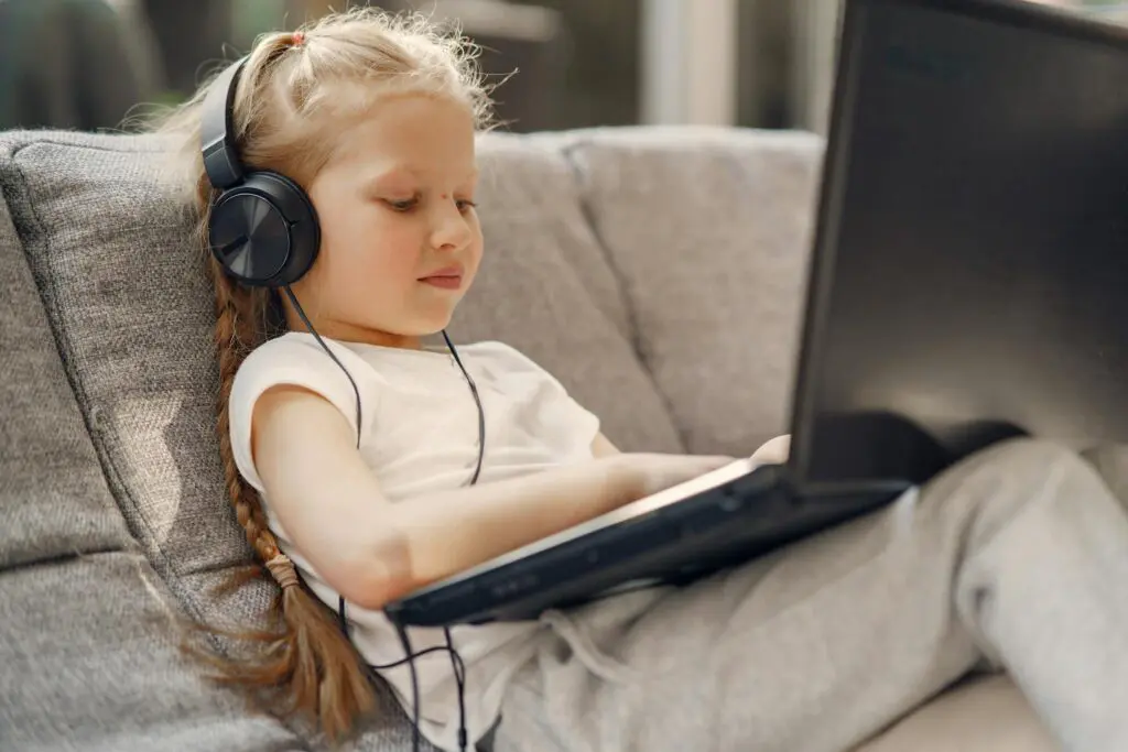 Small girl with long braid in headphones using laptop while sitting on sofa in living room