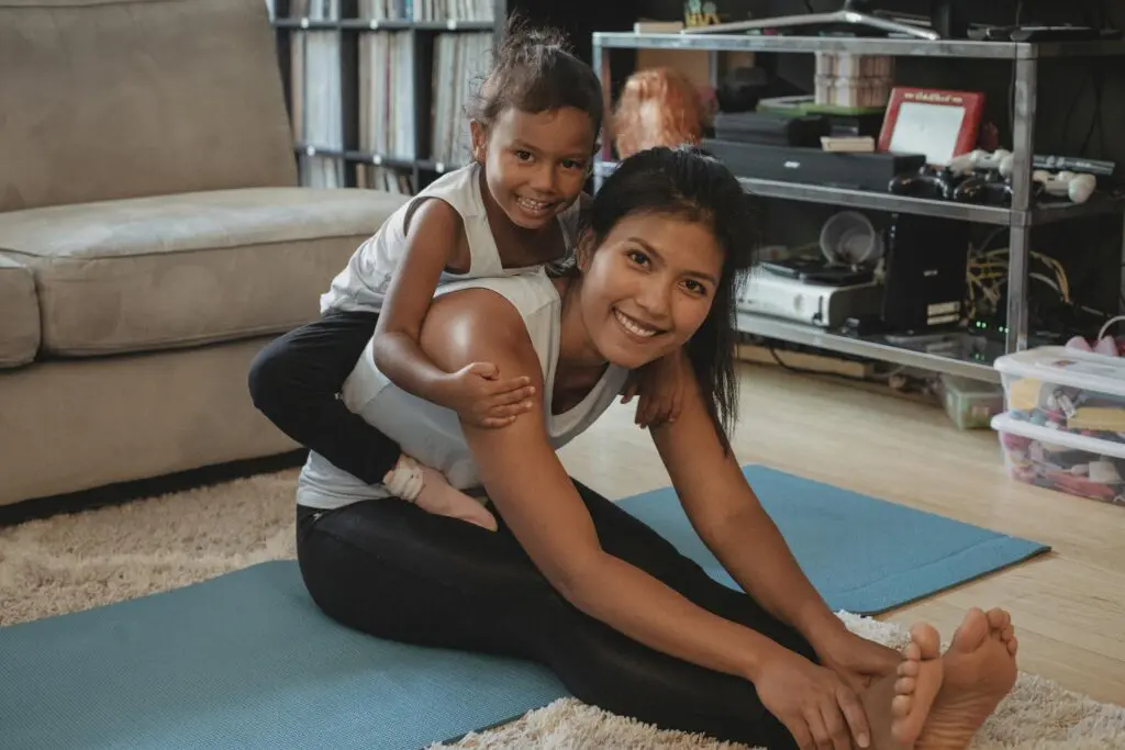 Happy ethnic mother stretching body and piggybacking adorable daughter while sitting on mat at home and looking at camera