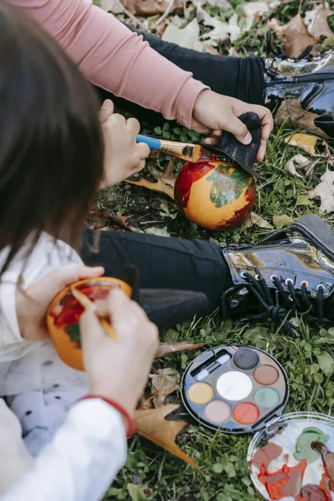 From above of crop anonymous little children sitting on grass and painting on small pumpkins